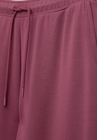 Detailní záběr na bordó tepláky, zobrazující texturu látky, postranní kapsu, elastický pas a šňůrku se plastovými koncovkami.