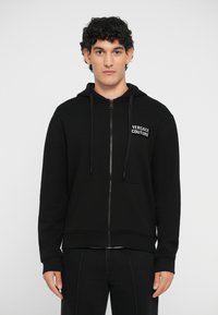 Mustmust värvi tõmbluku jack, mis on valmistatud puuvillast, esiPOOLKOTI ja joontega, ning millel on rinna peal "Versace Couture" logo.