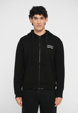 Musta vetoketjullinen huppari, joka on valmistettu puuvillamateriaalista. Siinä on edessä kengurulaukku, narut ja "Versace Couture" -logo rinnassa.