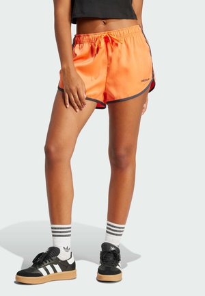 Pantaloni sportivi arancioni con rifiniture blu navy, vita elastica e stringa di regolazione. Indossati con sneakers nere e calze bianche con strisce nere.