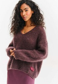 Sweter z dekoltem w serek w kolorze śliwkowym, wykonany z miękkiej, puszystej dzianiny, z luźnymi rękawami i detalem w postaci metki na dole. Nosi się go na fioletową spódnicę.