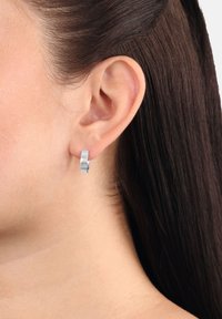 Kvinnas högra öra med rakt mörkbrunt hår som är nedstuckit bakom, bär en liten slät silverfärgad hoop-örhänge.