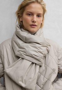 Beige Puffer-Schal mit genähtem Design, der eine weiche Textur und eine voluminöse Form aufweist und um einen hellen Rollkragenpullover gewickelt ist.
