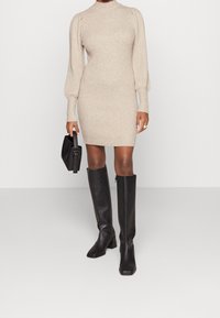 Abito in maglia a coste beige con colletto alto e maniche a sbuffo, abbinato a stivali neri sopra il ginocchio e una borsa nera strutturata.