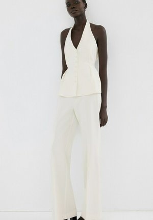 Donna dalla pelle scura che indossa un gilet bianco senza maniche con bottoni e pantaloni bianchi a gamba larga, in piedi davanti a uno sfondo bianco uniforme.