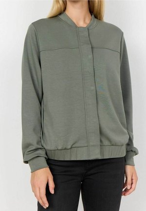 Femme portant une veste bomber vert olive à manches longues avec une fermeture éclair devant et une taille élastique, associée à un pantalon noir.