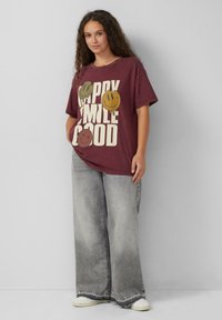 Bordowa, oversize koszulka z grafiką uśmiechniętej twarzy i białym napisem "HAPPY SMILE GOOD"; zestawiona z wyblakłymi szarymi spodniami typu wide-leg. Białe sneakersy.