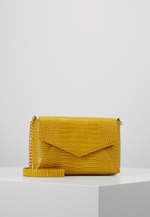 Żółta kopertówka ze skóry o fakturze krokodyla z złotym łańcuszkiem na białym pedestale na szarym tle.