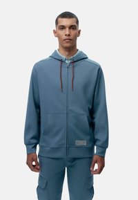 Zatvarač s kapuljačom u mekanom, laganom plavom materijalu. Ima kapuljaču s podesivom vrpcom, dva prednja džepa i malu oznaku s logotipom blizu rubnog šava.