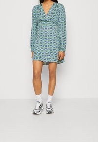 Mekko, jossa on pitkät hihat ja V-pääntie, sinivihreä geometrinen kuvio, keskipitkä pituus, vyötärö on istuva ja helma levenee.