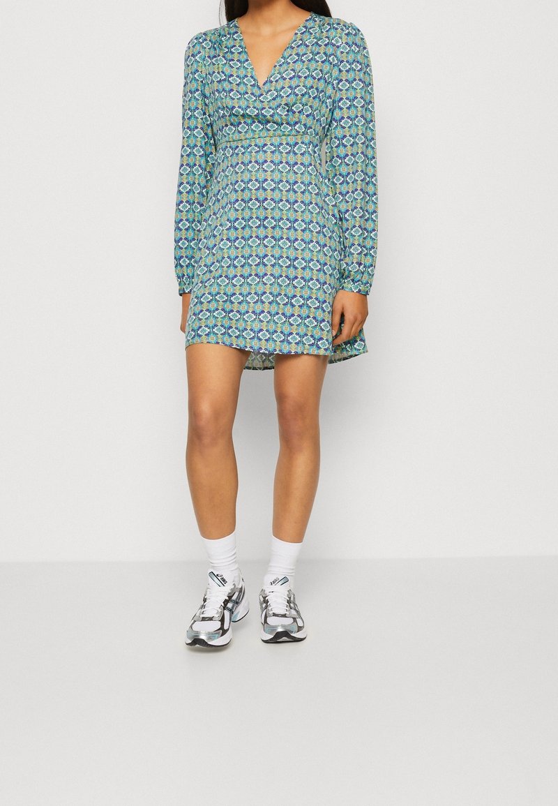 Šaty s dlouhým rukávem a výstřihem do V, s modro-zeleným geometrickým vzorem, do délky po stehna, se zdůrazněným pasem a rozšířeným lemem.