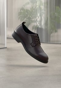 Καφέ δερμάτινο παπούτσι με ομαλή υφή, σχεδιασμό με κορδόνια, στρογγυλεμένη μύτη και επίπεδη ελαστική σόλα, τοποθετημένο στον αέρα με φόντο μινιμαλιστικό.