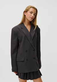 Blazer cinza escuro de dois botões feito de tecido texturizado, com lapelas recortadas, dois bolsos frontais e botões pretos. Saia plissada em camadas abaixo.