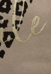 Tkanina bež boje s crnim uzorkom leoparda i sjajnim zlatnim rukopisnim natpisom. Teksturalne točke leoparda stvaraju kontrast s glatkom pozadinom.