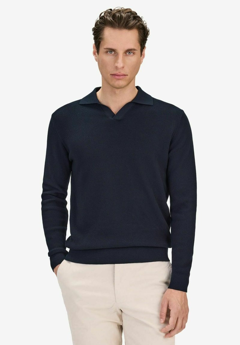 Homme portant un pull en maille bleu marine à manches longues avec col et un pantalon beige, debout avec une main dans la poche devant un fond uni.