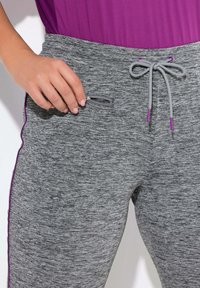 Graue Jogginghosen mit einem strukturierten Design, mit einer Reißverschlusstasche und lila Kordeln. Akzente sind lila Verzierungen entlang der Nähte.