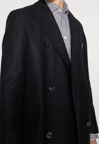 Cappotto nero doppiopetto realizzato in un tessuto testurizzato, caratterizzato da grandi bottoni lucidi e rever a punta, indossato sopra una camicia a righe.