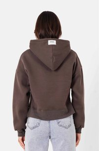 Sudadera corta marrón con capucha, puños y dobladillo acanalados. Etiqueta visible en la nuca; tela texturizada. Combinada con jeans de color azul claro.
