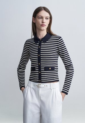 Donna che indossa un cardigan a righe blu navy e bianche con collo e bottoni, pantaloni bianchi a vita alta con cintura, mani nelle tasche, che guarda di lato.