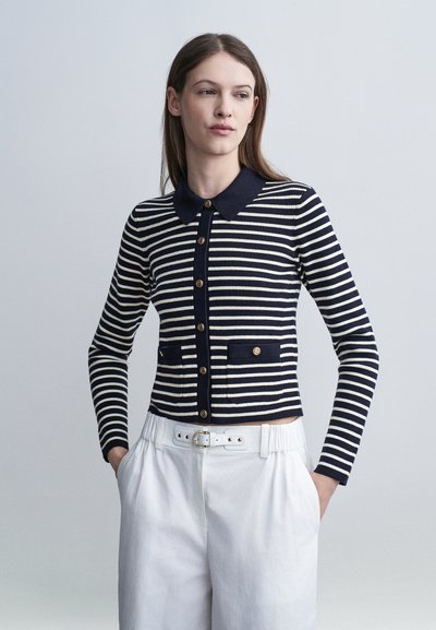 Femme portant un cardigan à boutons rayé bleu marine et blanc avec col, et un pantalon blanc taille haute avec ceinture, les mains dans les poches, regardant sur le côté.