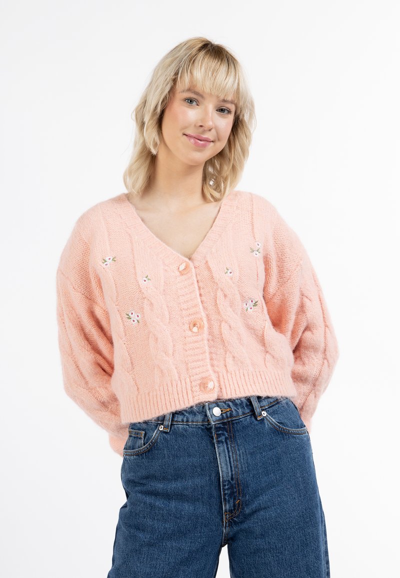 Cardigan a maglia rosa con scollo a V, grandi bottoni e dettagli floreali ricamati sul davanti. Abbinato a jeans a vita alta.