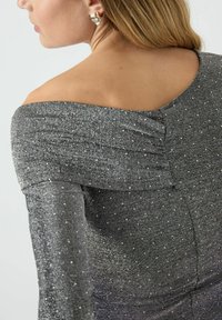 Grå metallic topp med avsvingte skuldre og teksturert stoff, med spredte sølvdetaljer og en innsvingt silhuett.