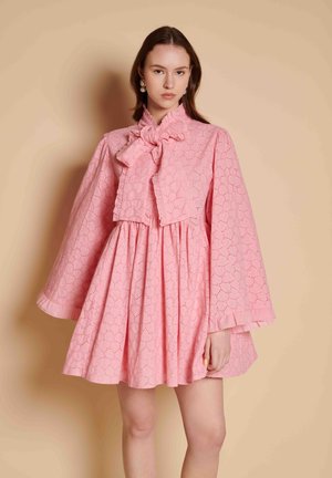 Young woman wearing a pink eyelet lace dress with a large bow at the neck and wide, long sleeves against a beige background.