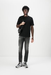 T-shirt noir, jeans gris foncé et baskets noir/blanc. Petit sac bandoulière noir avec une sangle. Le modèle se tient contre un fond blanc uni.