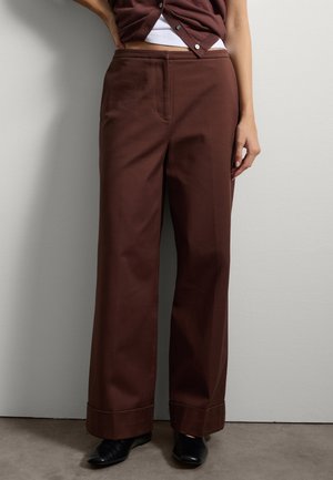 Pantalons larges marron en tissu robuste, avec des poches latérales et un ourlet à revers, associés à des chaussures noires sur un fond neutre.