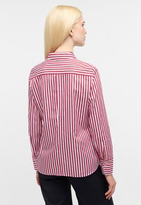Camicia a maniche lunghe a righe verticali rosse e bianche con colletto, caratterizzata da un orlo arrotondato e polsini con bottoni. Tessuto con una texture liscia.