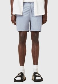 Pantaloni corti azzurri, lunghezza ginocchio, realizzati in tessuto morbido con vita elastica e cordoncino. Indossati con sandali neri e calzini bianchi.