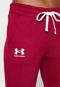 Burgunderfarbene Sporthose mit weißem Kordelzug und einem weißen Under Armour-Logo auf dem linken Oberschenkel. Weicher, strukturierter Stoff.
