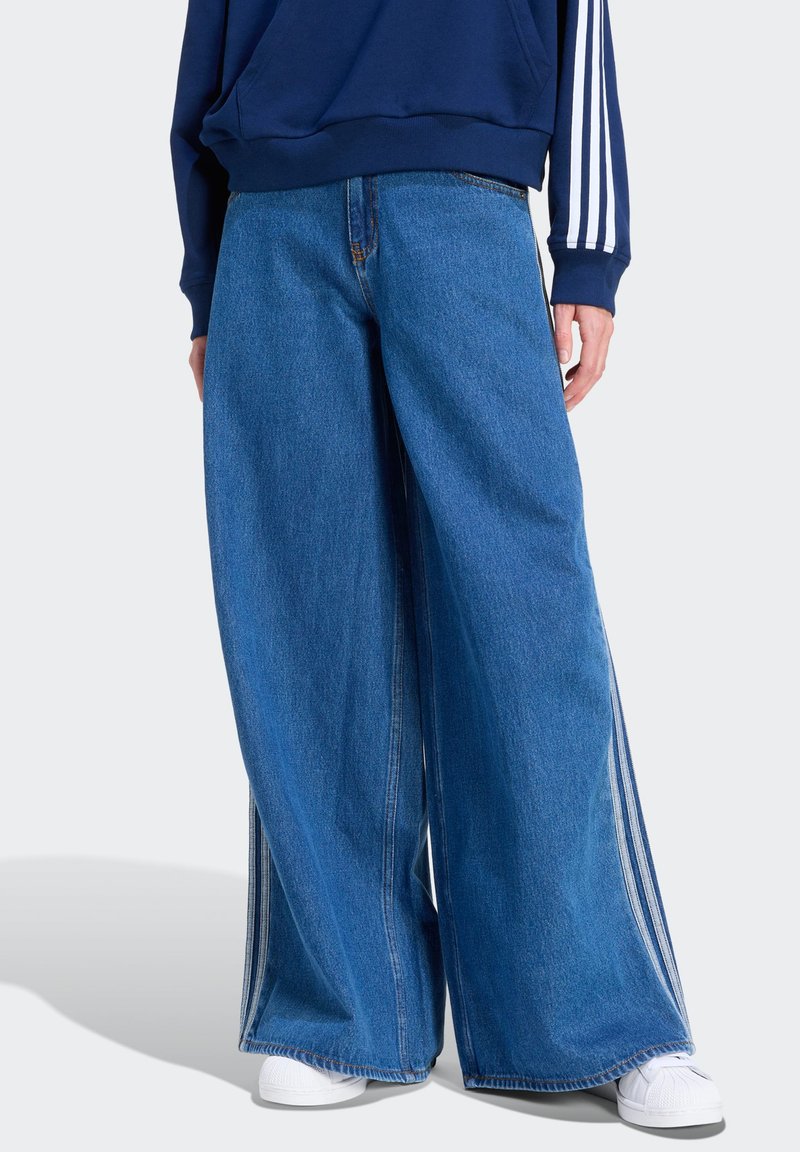 Jean en denim à jambes larges de couleur bleu clair avec une texture lisse, doté de bandes latérales blanches et de coutures contrastantes. Porté avec un sweat-shirt bleu marine.