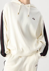 Cremfarbener Ripp-Hoodie mit schwarzen und roten Seitenstreifen, ausgestattet mit einer Kapuze mit Kordelzug und einem kleinen Logo auf der Brust. Weicher Stoff.