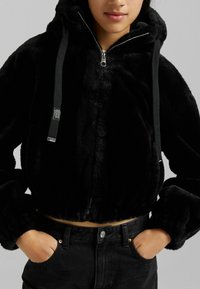 Felpa nera corta realizzata in morbido tessuto peluche. Presenta una zip frontale, un cappuccio regolabile con lacci, e polsini elasticizzati.