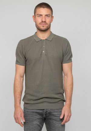 Homme aux cheveux courts et barbe, portant un polo à manches courtes vert olive et un jean gris foncé, debout devant un fond clair uni.