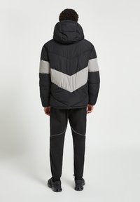 Giacca nera imbottita con strisce a zigzag grigie, cappuccio e polsini elastici. Abbinata a pantaloni neri con profili grigi e polsini elasticizzati alla caviglia.