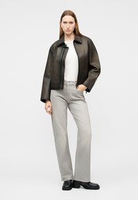 Γυναίκα που στέκεται φορώντας μαύρο δερμάτινο μπουφάν, λευκό πουκάμισο, ανοιχτό γκρι τζιν και μαύρα παπούτσια με πλατφόρμα, μπροστά από ένα απλό φόντο.