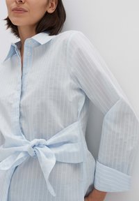 Camicia azzurra chiara con righe verticali bianche, design con colletto, maniche lunghe e dettaglio in vita annodato. Tessuto liscio con un aspetto fresco.