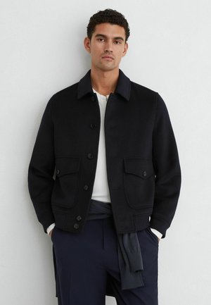 Uomo che indossa una giacca nera con bottoni sopra una camicia bianca e pantaloni scuri, in piedi contro un muro bianco semplice con le mani nelle tasche.