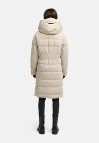 Beige gepolsterter langer Mantel mit tailliertem Schnitt und Kapuze, der eine glatte Textur und dezente Steppdetails aufweist. Schwarze Stiefel sind darunter sichtbar.
