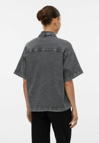 Camicia di jeans grigia con colletto corto, maniche corte e vestibilità rilassata. Tessuto trattato e cuciture visibili sulle giunture.