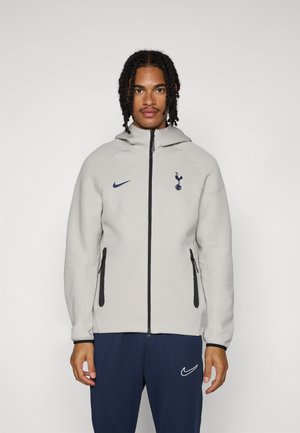 Light grey zip-up jacket with a hood. Features black accents, a Nike logo, and a Tottenham Hotspur emblem on the chest. Soft texture.