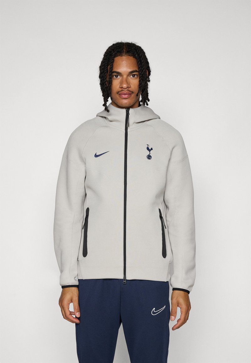 Light grey zip-up jacket with a hood. Features black accents, a Nike logo, and a Tottenham Hotspur emblem on the chest. Soft texture.