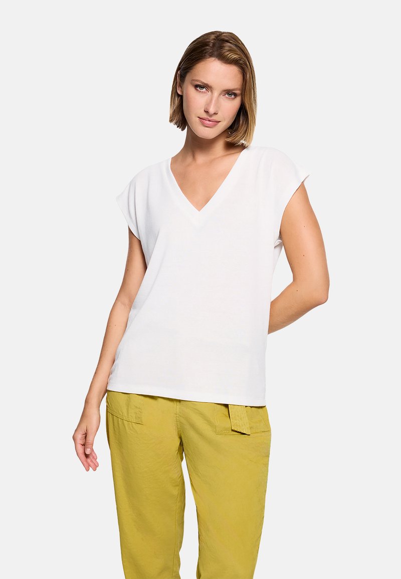 Mujer con cabello corto, vistiendo una camiseta blanca de manga corta con cuello en V y pantalones amarillos, de pie contra un fondo claro y sencillo.