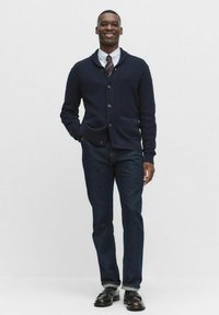 Marinblå cardigan med ribbad textur, knappar och två framfickor, parat med mörka jeans och svarta läderskor. Outfiten inkluderar en vit skjorta och slips.