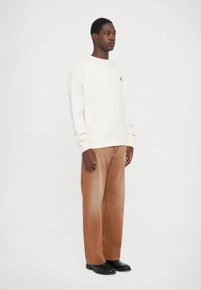 Mann som står iført en kremfarget genser med lange ermer, brune vidde jeans og svarte pensko mot en enkel hvit bakgrunn.