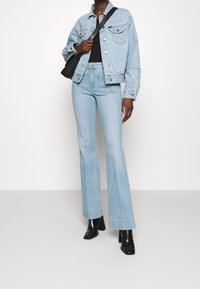 Hellblaues Jeansjackett mit silbernen Knöpfen und seitlichen Taschen, kombiniert mit passenden ausgestellten Jeans. Schwarze High Heels und eine Schultertasche.