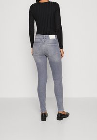 Jeans skinny grises con cintura de altura media, que cuentan con dos bolsillos traseros y una etiqueta blanca en el bolsillo derecho. El modelo lleva zapatos planos negros.
