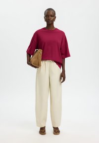 Γυναίκα στέκεται φορώντας μια άνετη σκούρα κόκκινη μπλούζα, κρεμ παντελόνι με φαρδιά μπατζάκια, ταμπά σανδάλια, κρατώντας ένα ταμπά σουέτ τσαντάκι κάτω από το χέρι της.